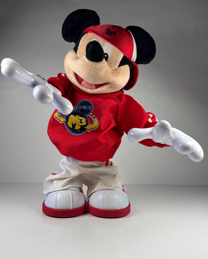 May include: A Mickey Mouse plush toy wearing a red and white outfit, including a red cap and sneakers. The red shirt has a logo with the letters "M3". The toy has white arms and is posed with arms outstretched.