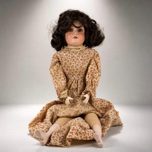 May include: A doll with dark brown curly hair and brown eyes, wearing a long-sleeved dress featuring a small red and yellow floral pattern on a beige background. The doll has light skin, visible arms, and legs with beige stockings.