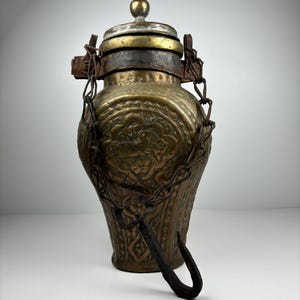 Puede incluir: Un recipiente antiguo de latón con intrincados patrones en relieve y una tapa con bisagras. El recipiente presenta una cadena y un asa curva. La tapa está rematada con un pequeño pomo redondo. El diseño sugiere su uso para líquidos o como objeto decorativo.