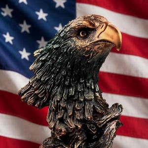 May include: A bronze-colored eagle sculpture with detailed feathering and a golden beak. The sculpture is set against a backdrop of the American flag, with its red and white stripes and blue field of stars.