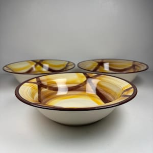 Puede incluir: Tres cuencos de cerámica con base crema y un patrón geométrico marrón y amarillo. Los cuencos tienen un borde ancho y poca profundidad, adecuados para servir ensaladas o guarniciones. Los cuencos están dispuestos en grupo.