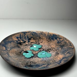 Puede incluir: Un plato decorativo redondo con una superficie moteada marrón y azul. Tres diseños de flores turquesas con centros blancos están dispuestos en el plato. El plato tiene un borde marrón oscuro.
