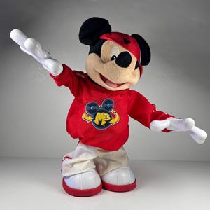 May include: A plush Mickey Mouse toy wearing a red sweatshirt with a yellow and blue graphic, white pants, and red shoes. The toy has black ears, a red hat, and white gloves. The toy is posed with its arms outstretched.