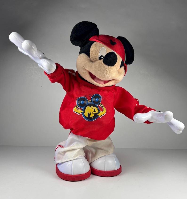 May include: A plush Mickey Mouse toy wearing a red sweatshirt with a yellow and blue graphic, white pants, and red shoes. The toy has black ears, a red hat, and white gloves. The toy is posed with its arms outstretched.