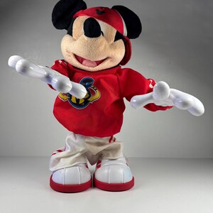 May include: A Mickey Mouse plush toy wearing a red baseball cap and a red long-sleeved shirt with a logo. The toy has white gloves and white and red sneakers. The plush is standing with its arms outstretched.