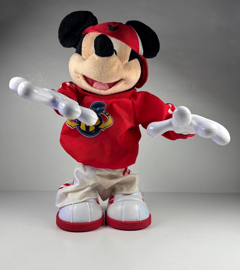 May include: A Mickey Mouse plush toy wearing a red baseball cap and a red long-sleeved shirt with a logo. The toy has white gloves and white and red sneakers. The plush is standing with its arms outstretched.