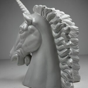 May include: A ceramic unicorn head sculpture in a matte white finish. The sculpture features a detailed mane, a spiraled horn, and a slightly open mouth. The unicorn is a mythical creature.