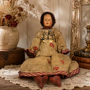 May include: An antique doll with a painted face and dark hair wearing a bonnet, a lace collar, and a tan dress featuring red floral patterns and a checkered hem. The doll has red painted feet and is dressed in a long-sleeved garment with a gold waistband.