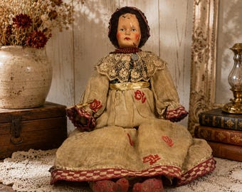 May include: An antique doll with a painted face and dark hair wearing a bonnet, a lace collar, and a tan dress featuring red floral patterns and a checkered hem. The doll has red painted feet and is dressed in a long-sleeved garment with a gold waistband.