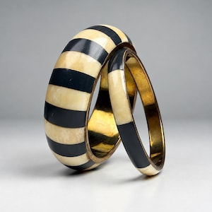 May include: Two stacked bangle bracelets featuring alternating black and cream-colored stripes. The wider bangle is positioned behind a thinner, matching bangle. Both bracelets have a smooth, polished finish and a metallic gold-toned interior.