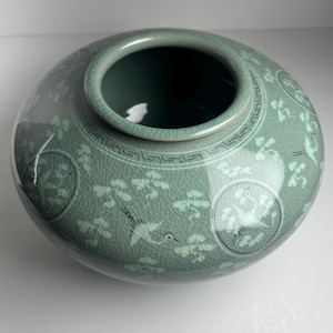 May include: A celadon ceramic vase with a wide, round body and a narrow opening. The vase has a crackle glaze and is decorated with floral and bird motifs. The design includes circular medallions and a decorative border. The vase is a pale green colour.