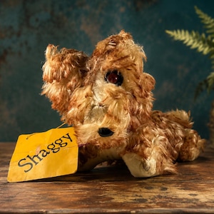 May include: A plush dog toy with long, shaggy brown fur, dark eyes, and a black nose. A yellow rectangular tag is attached to the toy, featuring the word Shaggy printed in black text. The toy is in a lying position with its head slightly raised.