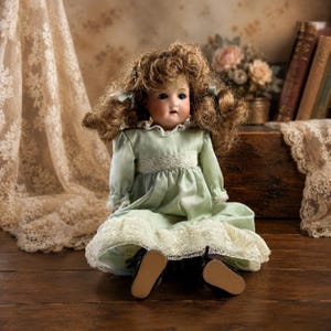 May include: A porcelain doll with curly brown hair and blue eyes, wearing a pale green dress with long sleeves, a high lace collar, a lace-trimmed waist, and a wide lace hem. The doll has dark brown shoes and is seated in an upright position.