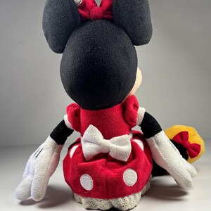 May include: A plush Minnie Mouse doll, viewed from the back, features a red dress with white polka dots and a white bow. The doll has black ears, a red bow with white polka dots, and white gloves. The doll is a classic Disney character.
