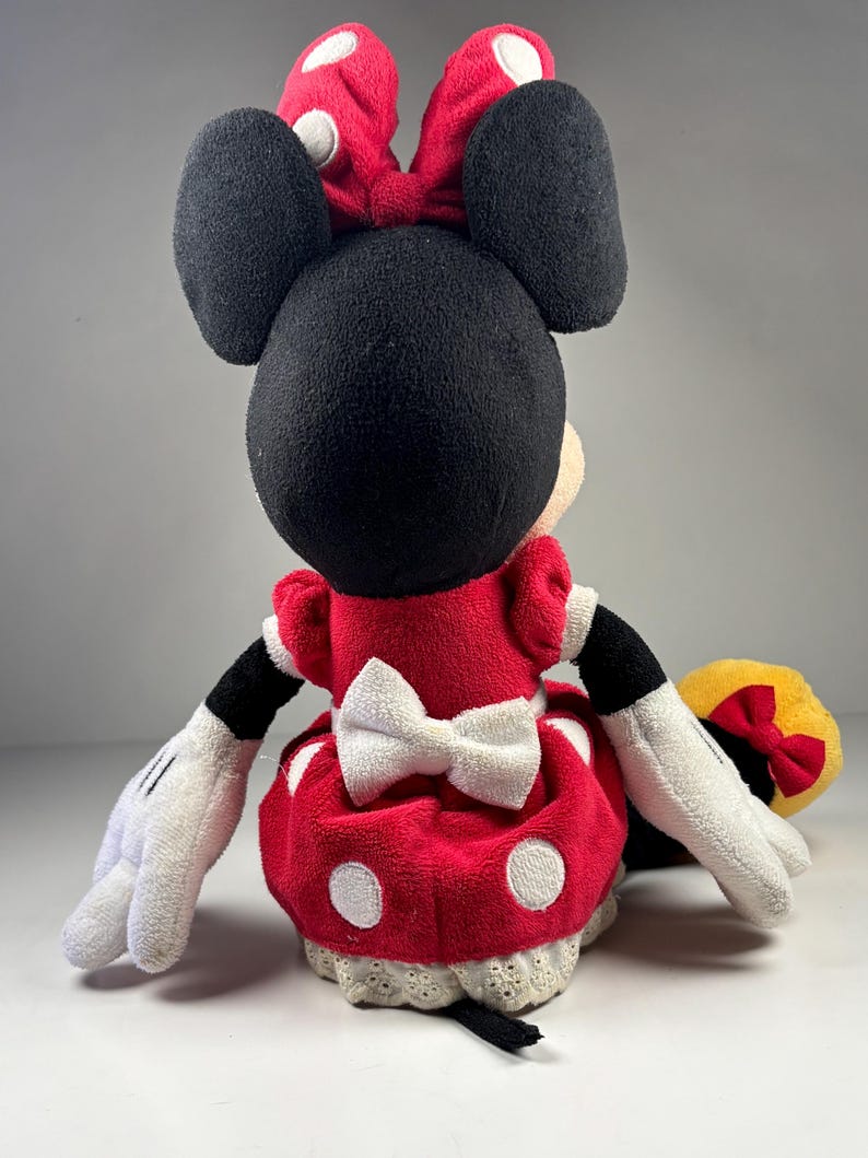 May include: A plush Minnie Mouse doll, viewed from the back, features a red dress with white polka dots and a white bow. The doll has black ears, a red bow with white polka dots, and white gloves. The doll is a classic Disney character.