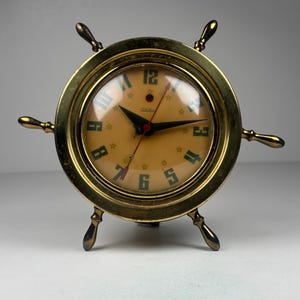 May include: A brass-colored clock shaped like a ship's wheel. The clock face is tan with black numerals and hands, and green star accents. The clock has four spokes and three feet.