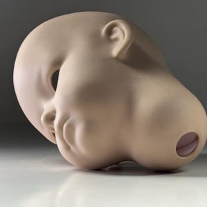 Op de afbeelding: Een bleke, gladde poppenkop met een gesloten mond, een duidelijke neus en een oor. De oogkas is een grote, open ovale vorm. De achterkant van het hoofd heeft een cirkelvormige opening. Het hoofd rust op een wit oppervlak.