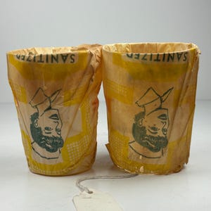 May include: Two yellow paper cups with a repeating yellow pattern and the word "SANITIZED" printed on the top. Each cup has a black and green illustration of a man's face. A blank tag hangs from the cups.