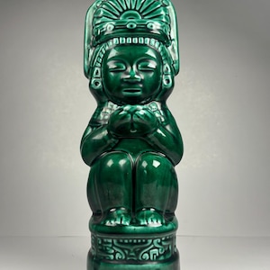 May include: A glossy, emerald green tiki mug depicting a seated figure with an elaborate headdress. The ceramic mug features a detailed design and the word "KAHECUA" at the base. The mug is a unique piece of barware.