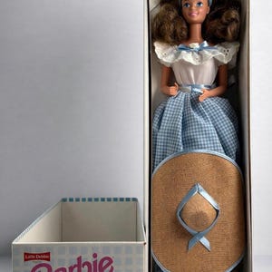 Op de afbeelding: Een Barbie Collector's Edition-pop in de originele doos. De pop heeft bruin haar en draagt een witte top met een blauw geruite rok en een strohoed. De doos heeft het Barbie-logo en de tekst "Collector's Edition Doll Series II."