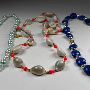 May include: Three necklaces of varying designs. One is a light blue pearl necklace, another features seashells and red beads, and the third is a necklace with blue stones and silver accents. The necklaces are displayed on a white surface.