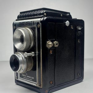 Puede incluir: Una cámara Spartaflex negra vintage con lentes y diales plateados. La cámara tiene un cuerpo texturizado y una placa de identificación metálica que dice "Spartaflex Camera Corp. Chicago, Ill. U.S.A."