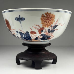 May include: A white porcelain bowl with floral and leaf designs in orange, red, and blue, resting on a dark wooden stand. The bowl's rim features a thin blue line. The artwork is reminiscent of traditional Asian pottery.