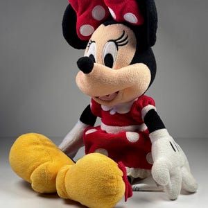 May include: A plush Minnie Mouse doll, featuring a red dress with white polka dots, a matching bow, and yellow shoes. The doll has black ears, a black nose, and white gloves with black details. The doll is sitting on a white surface.
