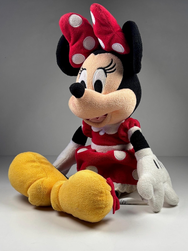 May include: A plush Minnie Mouse doll, featuring a red dress with white polka dots, a matching bow, and yellow shoes. The doll has black ears, a black nose, and white gloves with black details. The doll is sitting on a white surface.