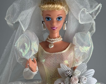 May include: A doll with blonde hair styled in an updo, wearing a white iridescent wedding gown with puff sleeves, a veil with small white fabric roses, a pearl necklace with a heart pendant, pearl drop earrings, and holding a white floral bouquet.