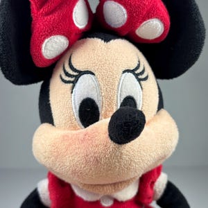May include: A plush Minnie Mouse doll with a red dress and bow. The doll has black ears, a black nose, and embroidered eyelashes. The red bow features white polka dots. The doll's dress has white trim and a white collar.