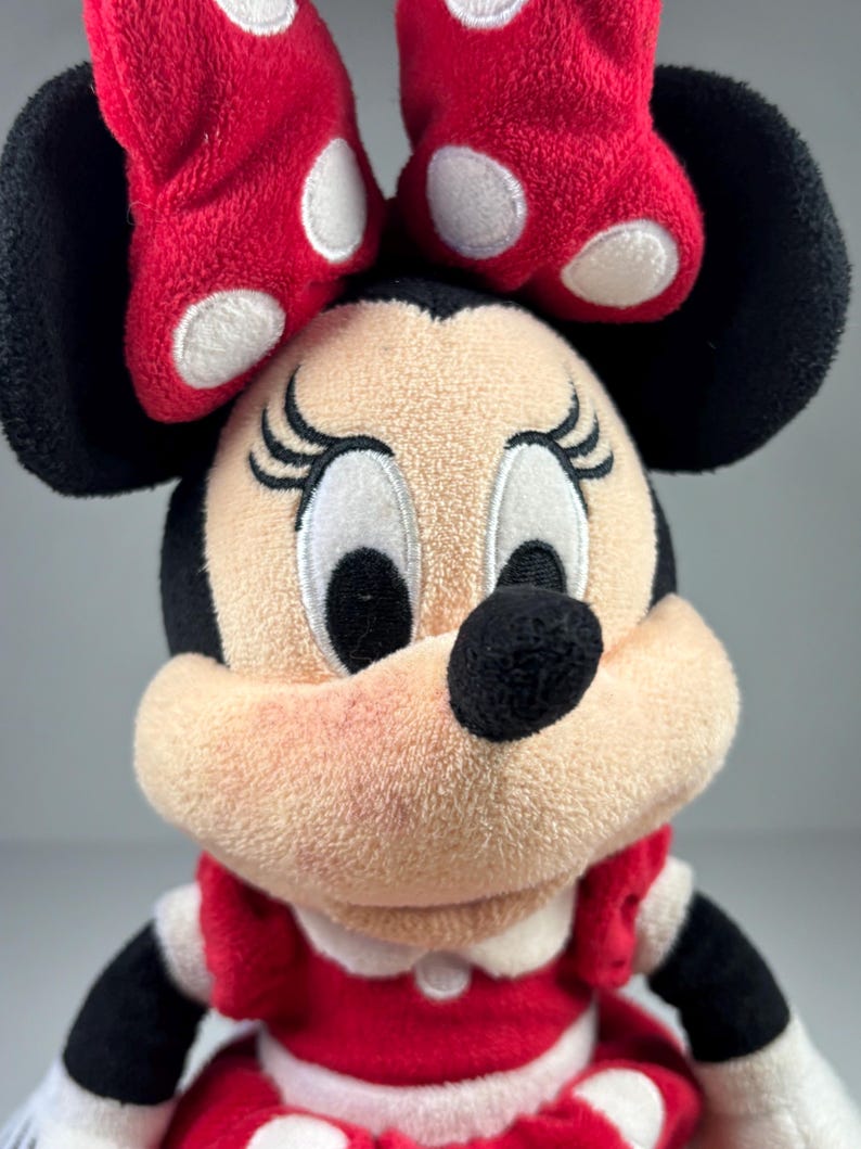 May include: A plush Minnie Mouse doll with a red dress and bow. The doll has black ears, a black nose, and embroidered eyelashes. The red bow features white polka dots. The doll's dress has white trim and a white collar.