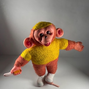 Puede incluir: Un juguete de peluche de mono vintage con un atuendo amarillo y naranja, zapatos blancos y una cara rosada. El mono sostiene un pequeño objeto rectangular en su mano derecha. El juguete tiene una expresión alegre y posa con los brazos extendidos.