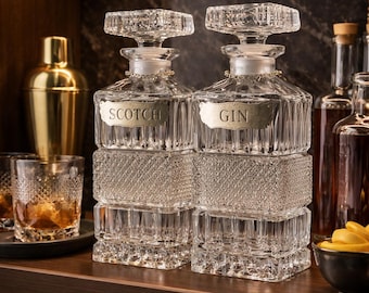 Op de afbeelding: Twee heldere glazen karaffen met vierkante bodem en gefacetteerde kristalpatronen. Elke karaf heeft een bijpassende glazen stop en een metalen label, de ene gegraveerd met SCOTCH en de andere met GIN.