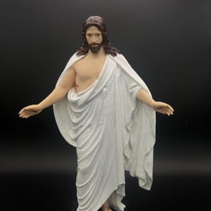 May include: A statue of a figure with long brown hair and a beard, wearing a white robe draped over the shoulders. The figure has outstretched arms and is standing on a black base. The statue is set against a black background.