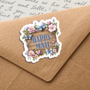 Op de afbeelding: Een sticker met de tekst "HAPPY MAIL" in blauw op een houten plank ontwerp, omringd door roze bloemen, blauwe vlinders en hartjes. De sticker is geplaatst op een bruine envelop.