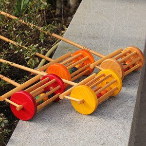 Könnte beinhalten: Vier hölzerne Spielzeug-Ziehwagen mit bunten Bällen im Inneren. Jeder Wagen hat ein Rad in einer anderen Farbe: rot, orange, gelb und grün. Die Wagen sind auf einer grauen Fläche aufgereiht.