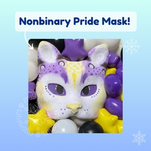 May include: A purple and yellow non-binary pride mask with a cat design. The mask is surrounded by purple, yellow, and white balls.