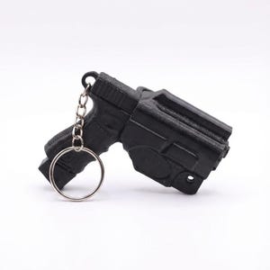 May include: Black keychain featuring a miniature pistol and holster. The gun is detailed with a textured surface and attached to a silver chain and ring. The keychain is a novelty item.