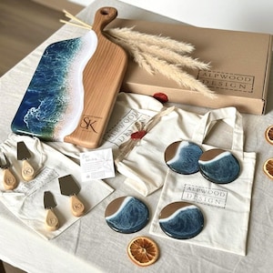 May include: A wooden serving board with blue and white resin ocean art, four round resin coasters, and four cheese knives with wooden handles. The set includes branded cotton drawstring bags, a cardboard box labelled ALPWOOD DESIGN, and dried orange slices.