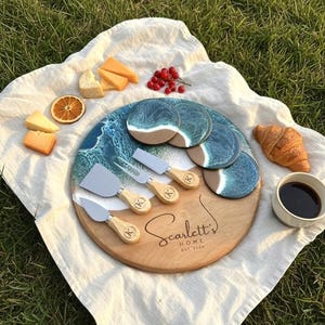 Może przedstawiać: Drewniana deska do serów z motywem fal, nożami do sera i pasującymi podstawkami. Deska jest grawerowana napisem "Scarlett's HOME EST. 2024". Scena zawiera ser, jagody, plaster pomarańczy, rogalik i filiżankę kawy.