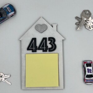 May include: A white wooden house-shaped sign with a silver heart and the number 443. A yellow sticky note is attached to the sign.