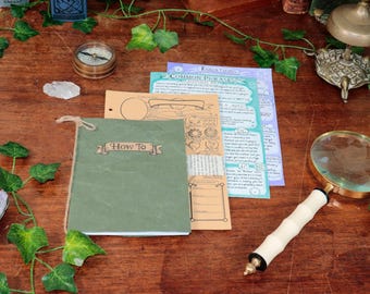 May include: A collection of items on a wooden surface, including a green notebook with "How To" text, a magnifying glass, and printed papers. A compass, crystals, and decorative ivy are also present.