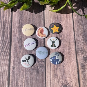 May include: Eight round pins with various designs. The pins feature a dragon, a deer, a crescent moon, a star, a mountain, and the words "Velaris", "Terrassen", and "Adarlan".