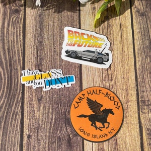 K&ouml;nnte beinhalten: Drei Aufkleber auf einem Holzgrund. Die Aufkleber sind ein runder orangefarbener Aufkleber mit einem schwarzen Pegasus und dem Text "Camp Half-Blood Long Island, N.Y.", ein blauer und gelber Aufkleber mit dem Text "Ich habe Unterricht und du nicht", und ein wei&szlig;er Aufkleber mit einem silbernen Auto und dem Text "Zur&uuml;ck in die Zukunft".