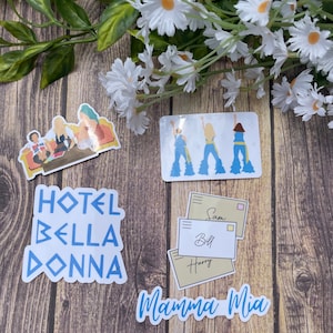 May include: A set of four stickers featuring characters and text from the movie Mamma Mia! The stickers are on a wooden surface. The stickers include a group of three women in blue dresses, a group of three women in a hotel room, a set of three envelopes with names on them, and the text "Mamma Mia!" in blue letters.