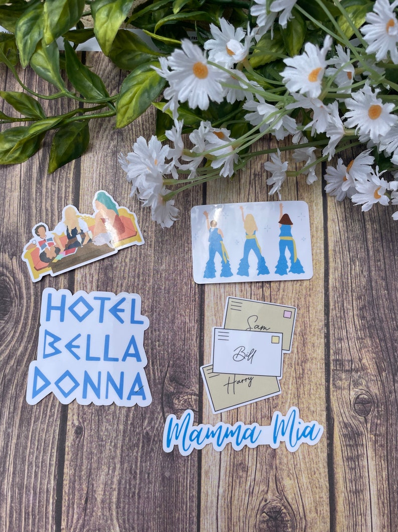 May include: A set of four stickers featuring characters and text from the movie Mamma Mia! The stickers are on a wooden surface. The stickers include a group of three women in blue dresses, a group of three women in a hotel room, a set of three envelopes with names on them, and the text "Mamma Mia!" in blue letters.