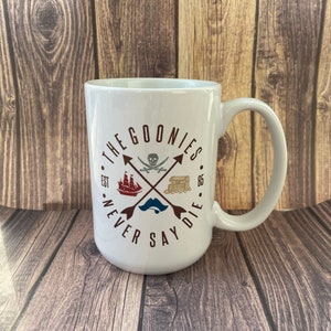May include: White ceramic mug with a brown graphic of a skull and crossbones with the text "The Goonies Est. '85 Never Say Die" printed on the front.