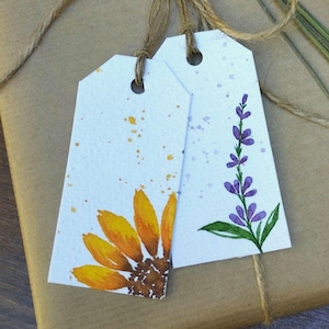 Op de afbeelding: Een cadeau verpakt in bruin papier, vastgebonden met touw en versierd met gedroogde lavendel en twee decoratieve labels. Eén label toont een aquarel zonnebloem, de andere een lavendel tak. Het cadeau wordt gepresenteerd op een houten oppervlak.