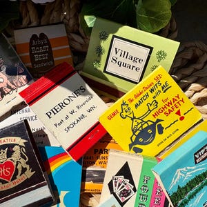 May include: A collection of vintage matchbooks with various designs and colors. The matchbooks feature text and graphics for restaurants, hotels, and other businesses. Some matchbooks have a red, white, and black color scheme, while others are blue, green, and yellow.